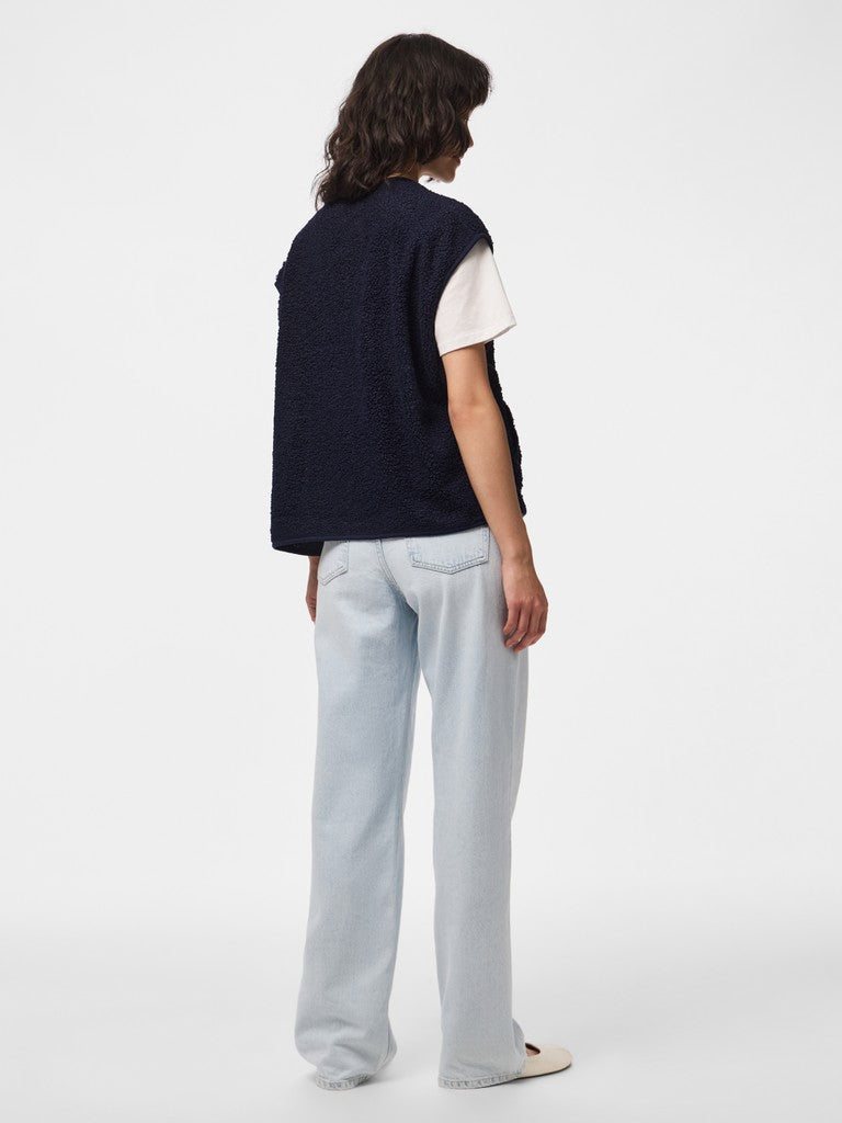 A model shown from the back wearing a dark navy sleeveless knit vest over a white t-shirt, paired with light-wash wide-leg jeans and white slip-on shoes, against a plain white studio background.