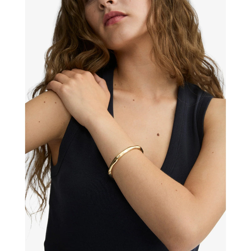 A woman wearing a slim, polished gold bangle bracelet on her wrist, paired with a dark navy sleeveless top against a clean white background.