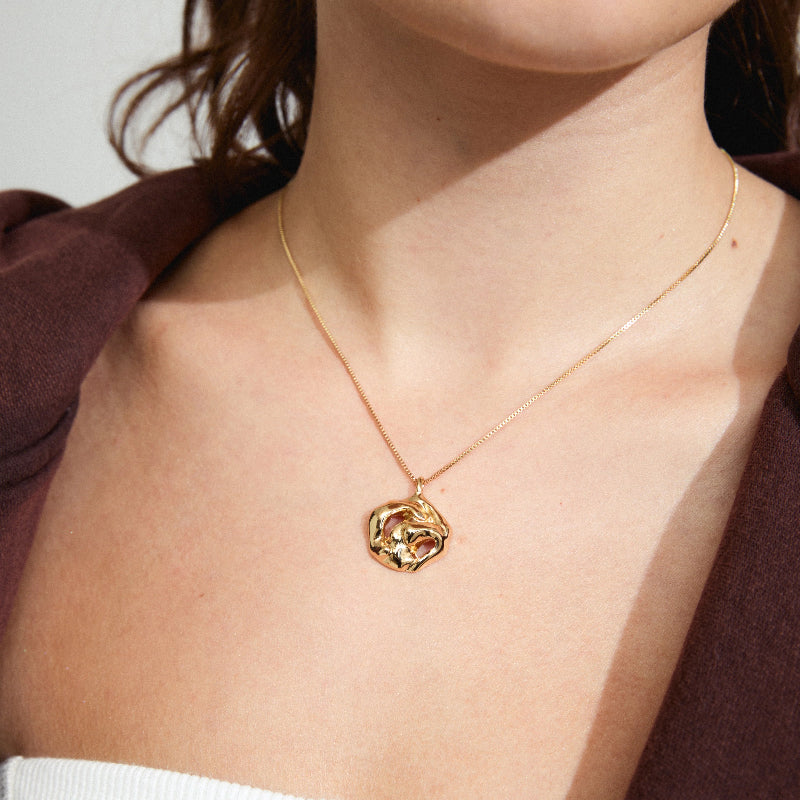 A close-up of a model wearing a delicate gold chain necklace with an organic, textured gold pendant.