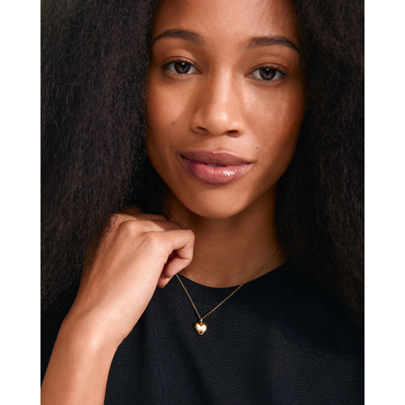 Woman wearing a gold necklace with a heart pendant against a neutral background