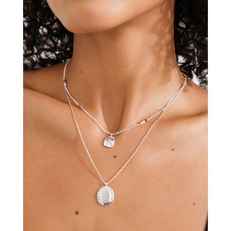 A close-up of a model's neck and chest wearing a layered silver necklace set. The top layer is a delicate chain with small colorful beads and a clear faceted pendant, while the bottom layer is a slightly thicker chain with a larger oval pendant featuring a silver filigree design.