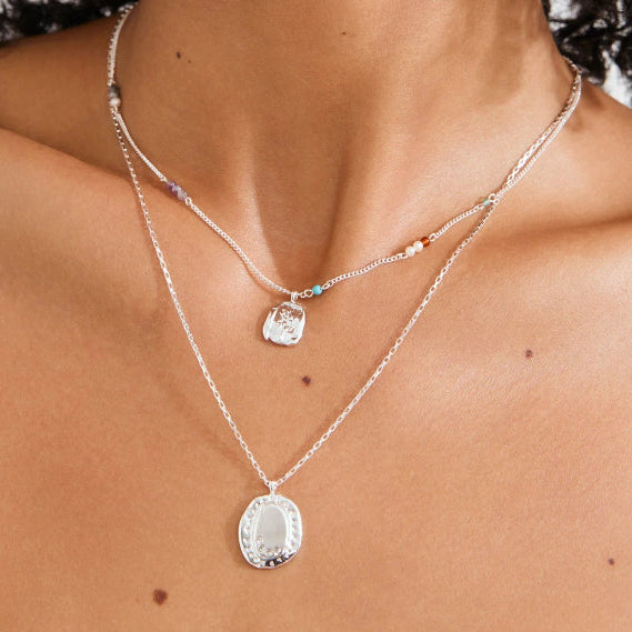 A close-up of a model's neck and chest wearing a layered silver necklace set. The top layer is a delicate chain with small colorful beads and a clear faceted pendant, while the bottom layer is a slightly thicker chain with a larger oval pendant featuring a silver filigree design.