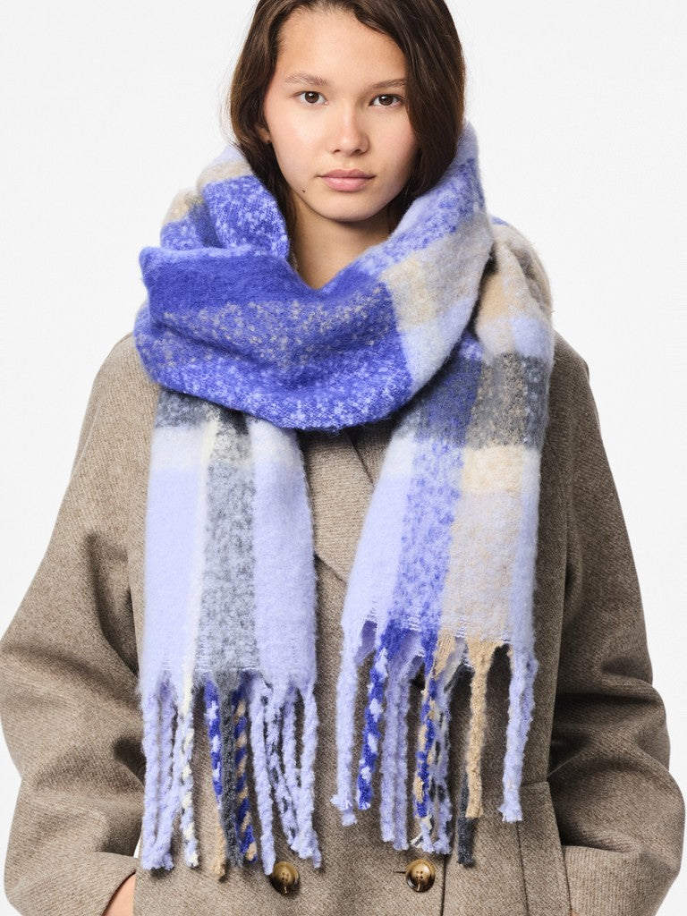 A woman wearing a thick, fuzzy plaid scarf in shades of blue, lavender, and beige, paired with a brown coat against a plain white background.