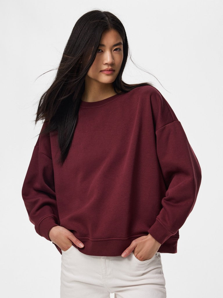 A woman wearing an oversized crewneck sweatshirt in deep burgundy, paired with white pants, photographed against a clean white studio background.