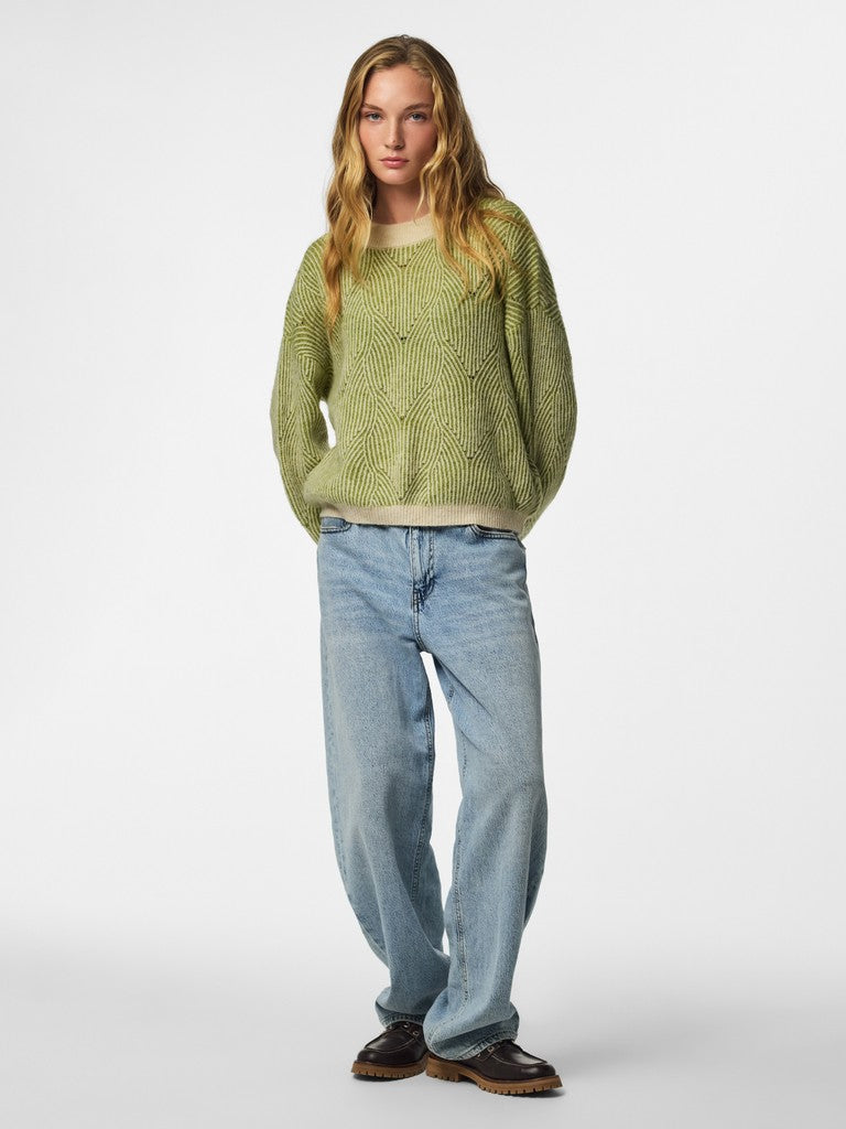 A woman modeling a light olive green textured knit sweater, paired with light-wash blue jeans and dark brown shoes, against a plain light gray studio background.