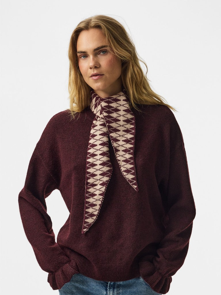 A woman wearing a dark burgundy knit sweater and a matching burgundy and cream patterned knit scarf, photographed against a light gray studio background.