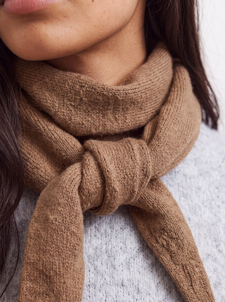 A close-up of a person wearing a chunky knit brown scarf tied around the neck over a light gray knit sweater.