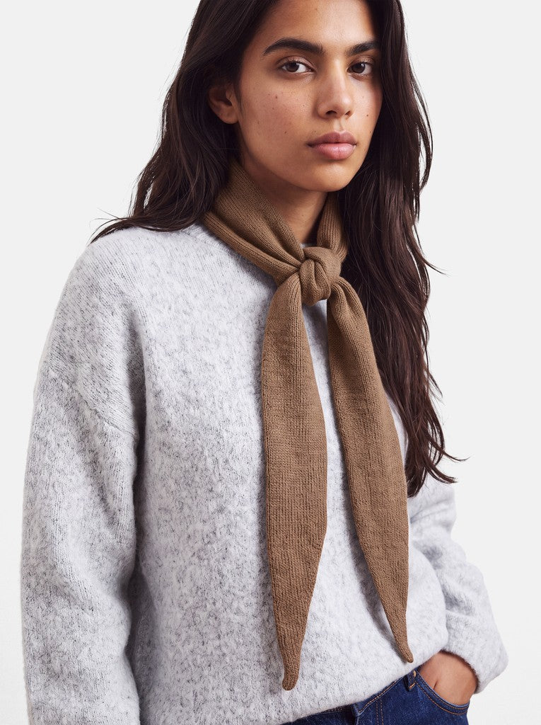 A woman wearing a light gray knit sweater and a brown knotted scarf, photographed against a clean off-white studio background.