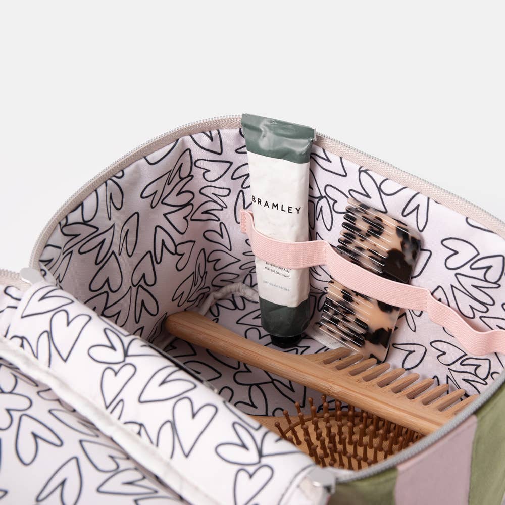 An open zippered toiletry bag with a white interior printed with black heart outlines and a pink elastic organizer. Visible items inside include a tube labeled Bramley, a leopard-print hairbrush, a wooden comb, and a brown bristle brush.