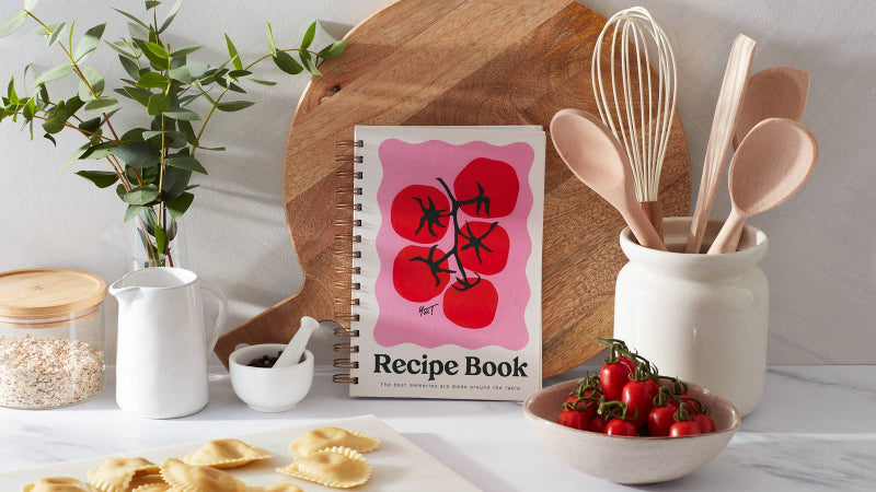 Recipe book with red tomato design on a kitchen counter with utensils and ingredients.