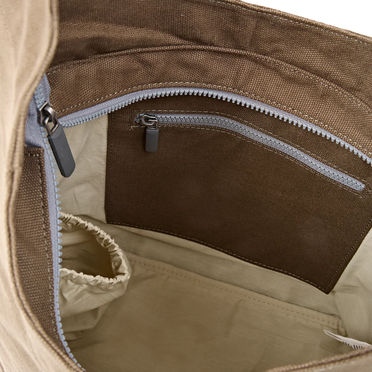 Close-up of the interior of a brown canvas bag, showing a zippered pocket with a gray zipper pull and a light beige fabric lining.