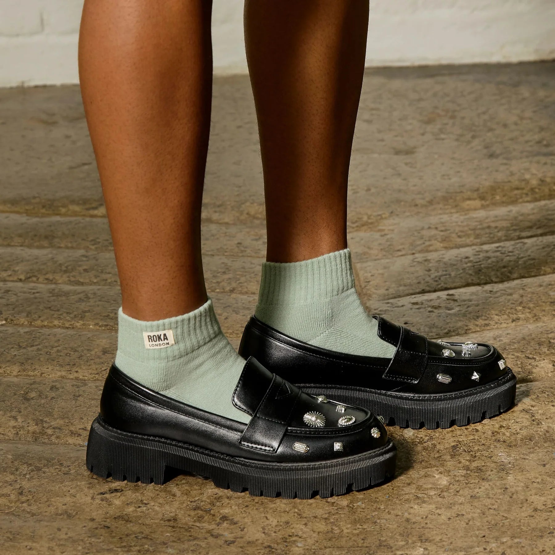 Black loafers with studded details worn with light green socks on a wooden floor.