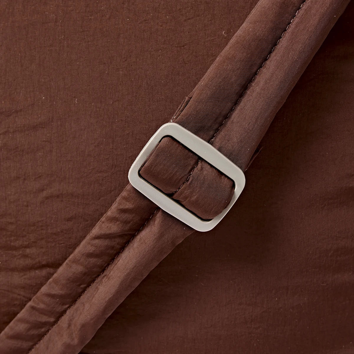 Close-up of a brown fabric strap with a silver-toned rectangular buckle, set against matching brown textured fabric.