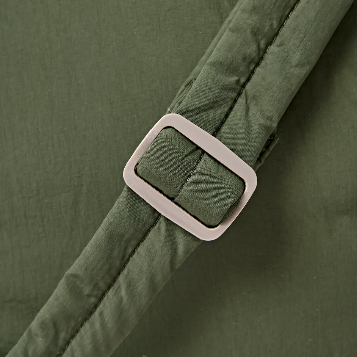 Close-up of an olive green padded strap with a matte light gray rectangular buckle, shown against matching olive green fabric.