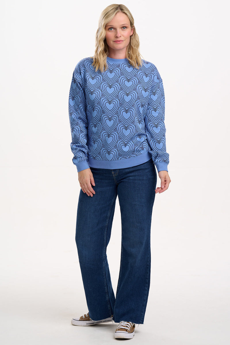 Woman wearing a Sugarhill Blue Heart Explosion Sweatshirt and blue jeans on a white background