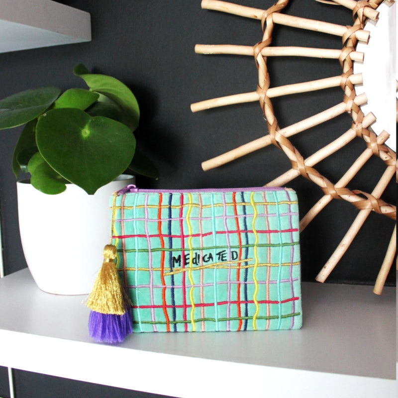 A colorful embroidered cosmetic pouch with a purple and mint green plaid pattern, a gold tassel, and the text 'MEDICATED' on the front, displayed on a white shelf next to a potted plant and a wooden sunburst wall decor.