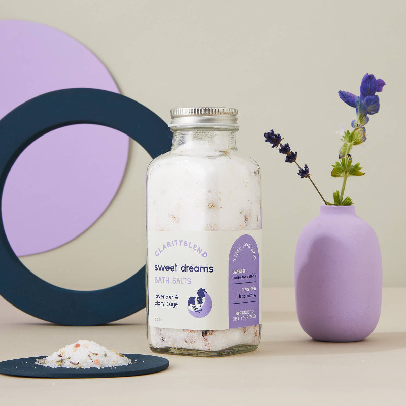 A glass jar of Clarity Blend Sweet Dreams Bath Salts with lavender and clove oil, styled on a beige surface with a small pile of salts in a dark blue dish, a purple ceramic vase with flowers, and abstract geometric shapes in the background.