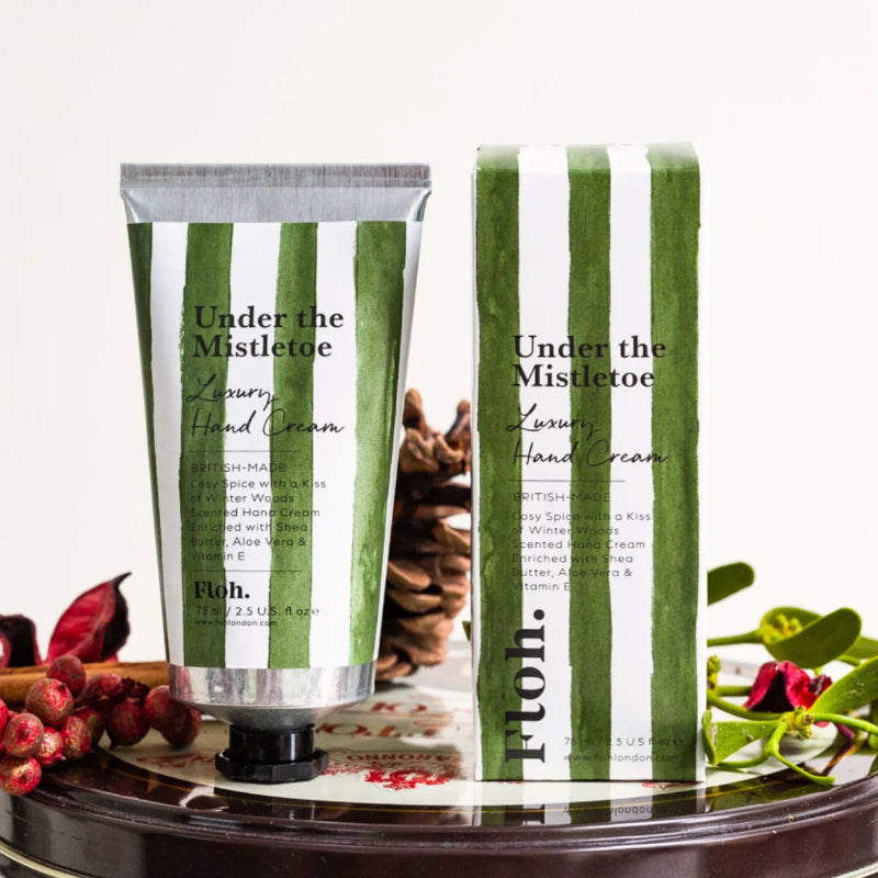 A squeeze tube and matching box of Floh. 'Under the Mistletoe' Luxury Hand Cream, decorated with green and white vertical stripes, displayed on a dark reflective tray with pinecones and red berries.
