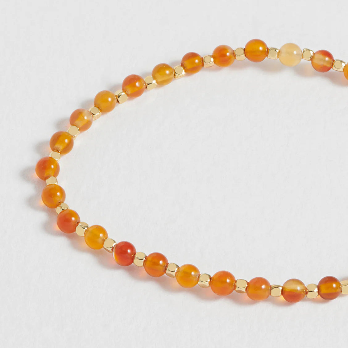 A beaded strand necklace or bracelet made of round orange and amber gemstone beads separated by small gold spacer beads, photographed on a plain light background.