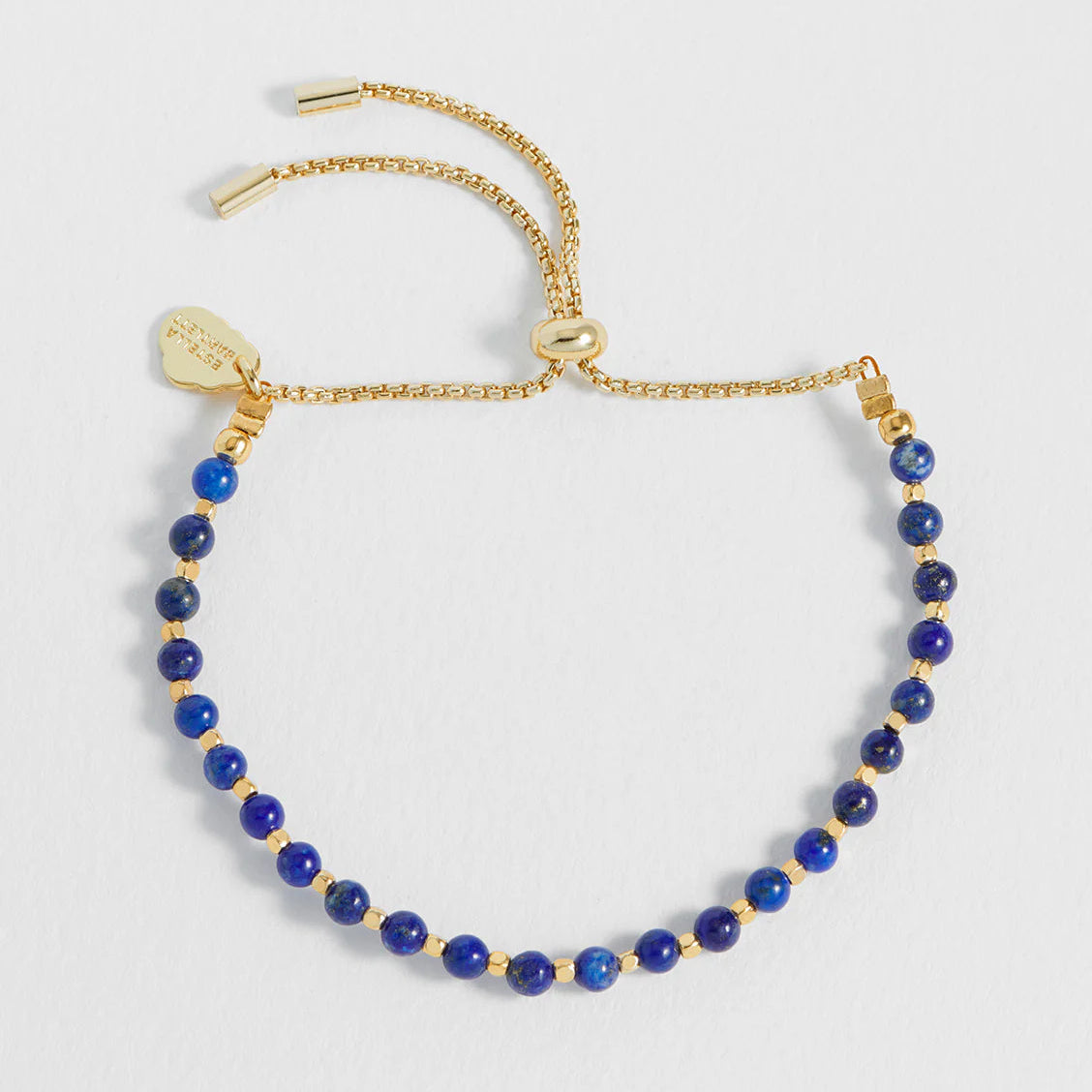 A Estella Bartlett gold-tone adjustable chain bracelet featuring a row of round, deep blue lapis lazuli-style beads separated by small gold spacer beads, with a small heart-shaped charm and sliding clasp.
