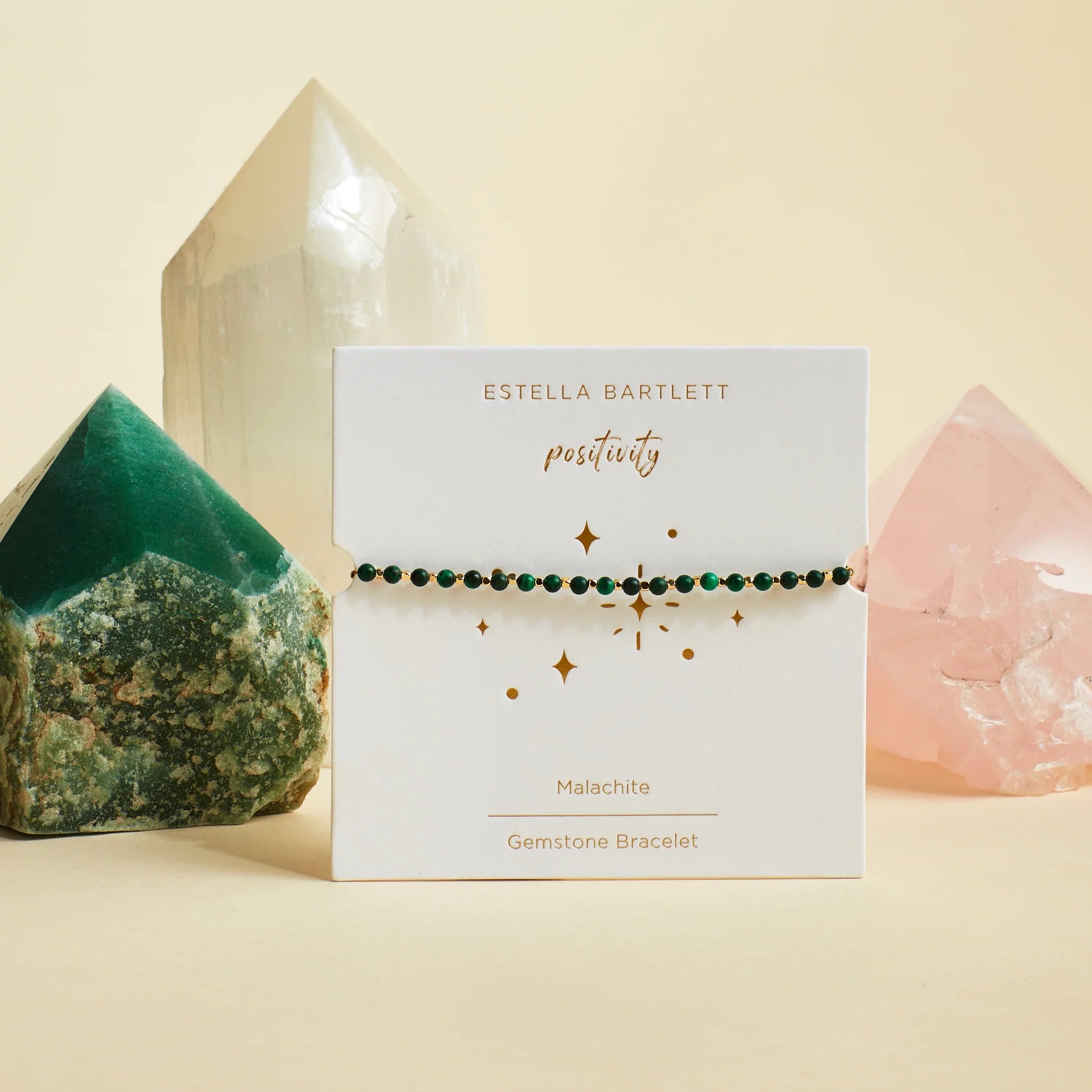 A delicate green gemstone bracelet by Estella Bartlett, displayed on its white branded card. The card is styled on a beige surface with crystal points in the background.