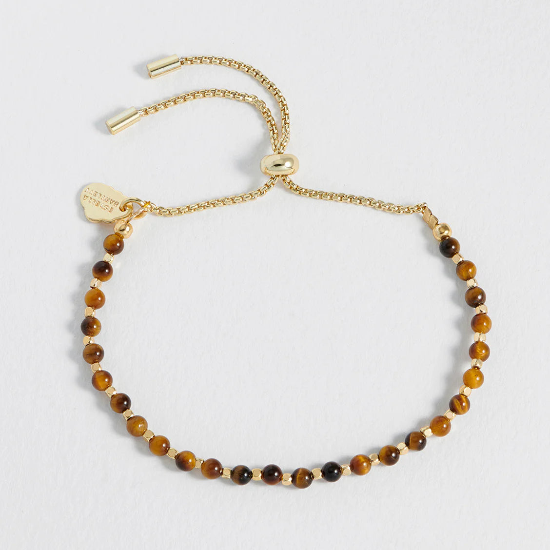 A delicate gold chain bracelet with small brown and black tiger's eye beads, featuring an adjustable sliding clasp and a small heart-shaped tag, displayed on a light gray background.