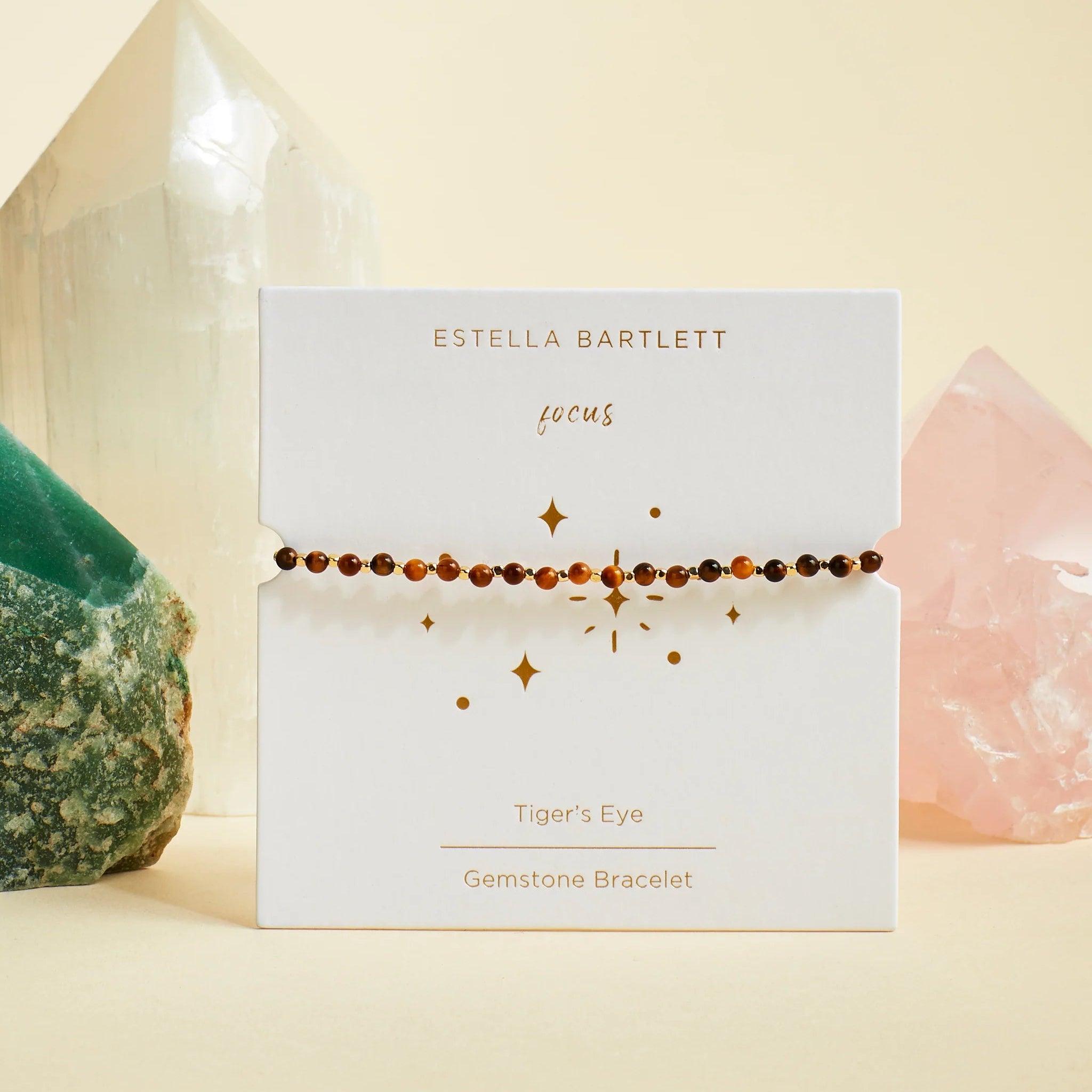 A tiger's eye gemstone bracelet by Estella Bartlett, displayed on its white branded card packaging. The bracelet consists of small brown tiger's eye beads strung across the card.