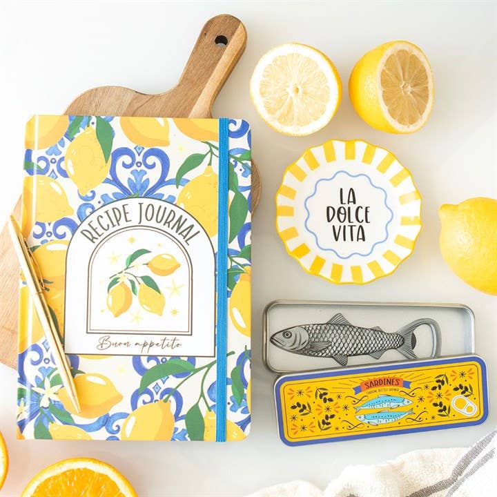 Recipe journal with lemon design, lemons, and a 'La Dolce Vita' plate on a white surface.
