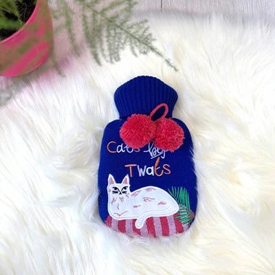 A navy blue knitted hot water bottle cover with a white cat illustration and the text 'Cats Love Twats' in orange and white lettering. It features two red pom-poms on the neck and is displayed on a white faux fur surface.