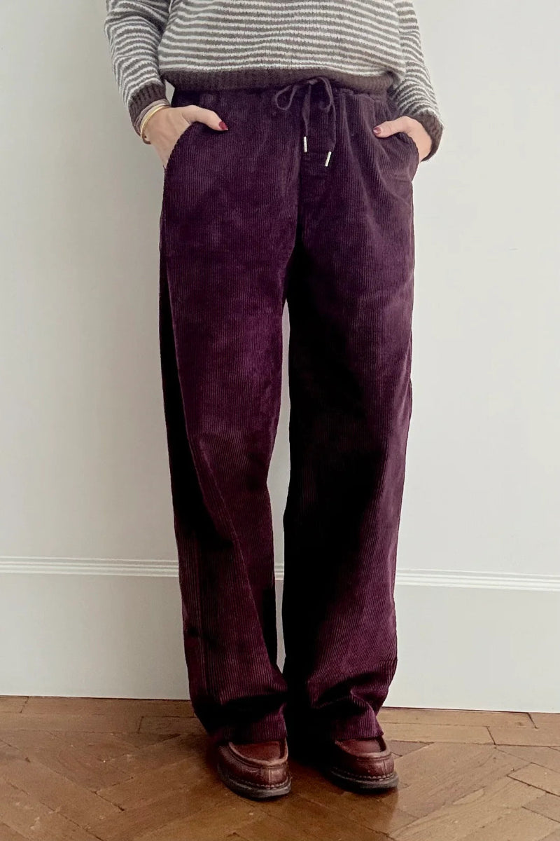 Person wearing purple corduroy pants and a striped sweater against a white wall.