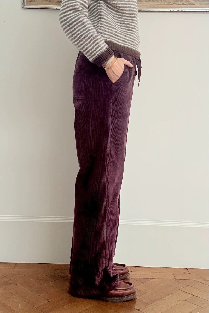 Person wearing purple velvet pants and a striped sweater against a white wall.