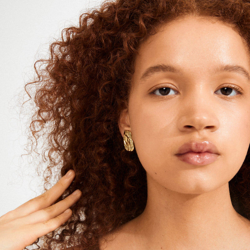 Close-up of a person with curly hair wearing Pilgrim Shift Earrings Gold-plated against a white background