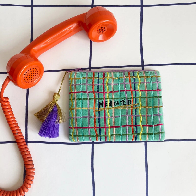A green plaid fabric pouch with a gold zipper and a purple tassel charm, featuring the text 'MEDCATED' on the front. It is styled on a white grid background next to a retro orange rotary telephone receiver.