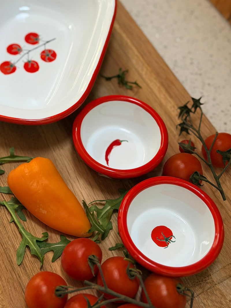 Set of ceramic dishes with red rim and tomato design on a wooden cutting board with vegetables.