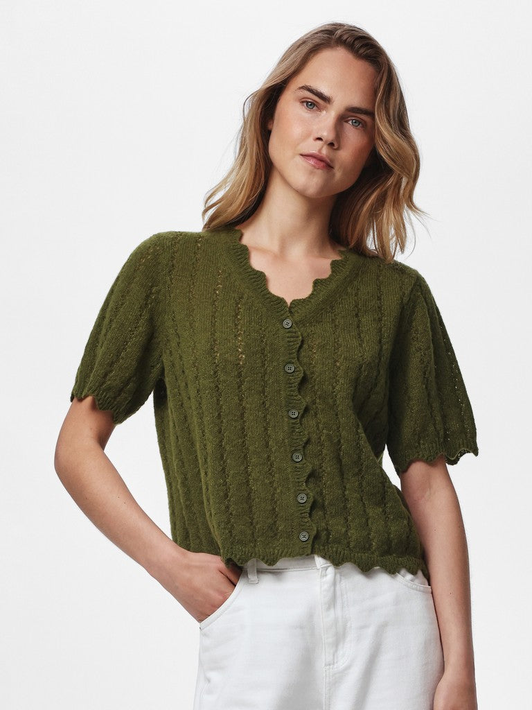 A woman wearing a short-sleeve olive green knit top with a scalloped neckline and front buttons, paired with white pants, against a plain light gray studio background.