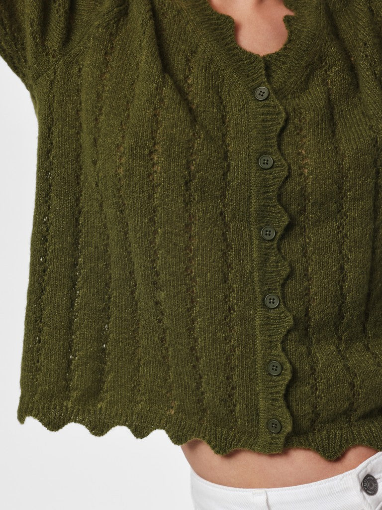 Close-up of a person wearing an olive green ribbed knit cardigan with a scalloped edge and dark buttons, paired with white pants.