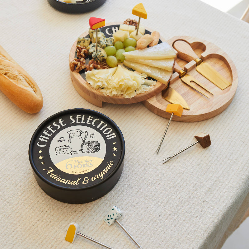 A styled cheese board setup on a cream tablecloth, featuring a round black and white 'Cheese Selection' tin, a wooden serving board with assorted cheeses, crackers, grapes, and cheese knives, and several cheese picks with small dice and yellow-tipped skewers.