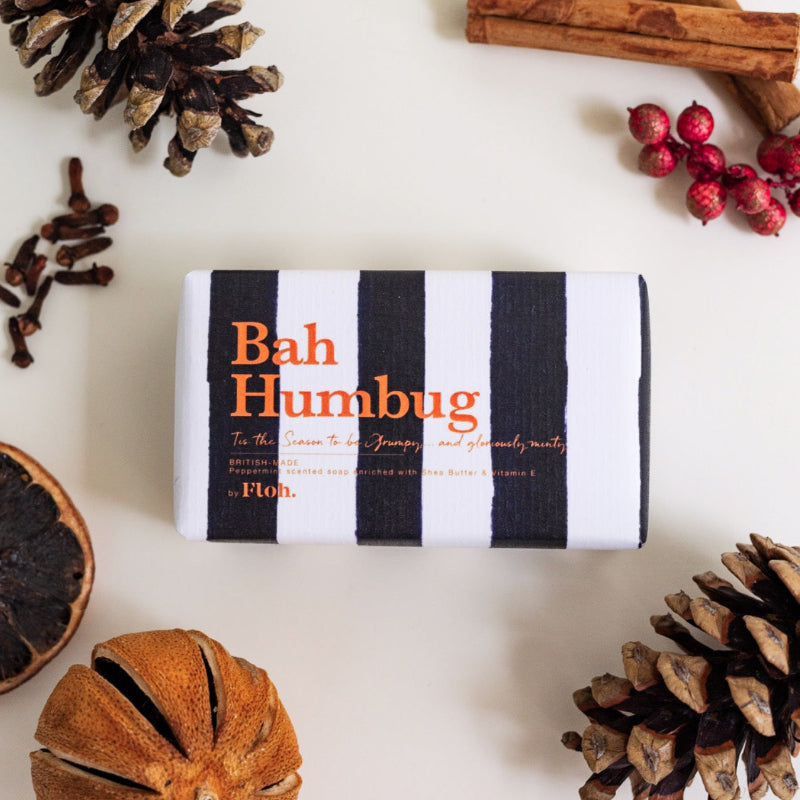 A wrapped bar of soap labeled 'Bah Humbug' by Flo. is arranged in a flat lay on a white surface with seasonal props including pinecones, cloves, cranberries, cinnamon, and a dried orange slice.
