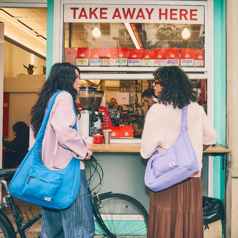 Two women with Roka Belize shoulder bags standing in front of a 'Take Away Here' sign at a food stand.