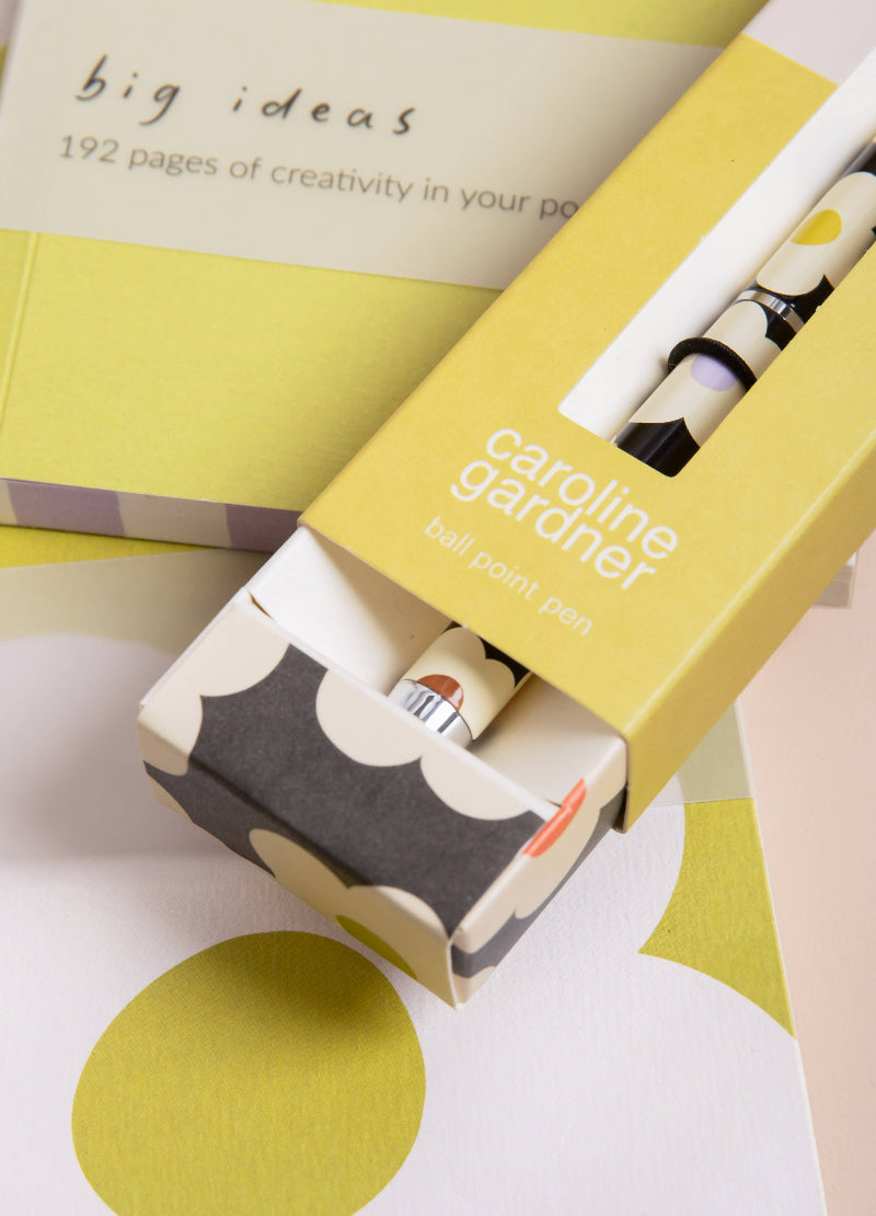 A Caroline Gardner ball point pen in a yellow and gray patterned box, shown alongside a matching book titled 'big ideas' on a white surface with large yellow and white abstract shapes.