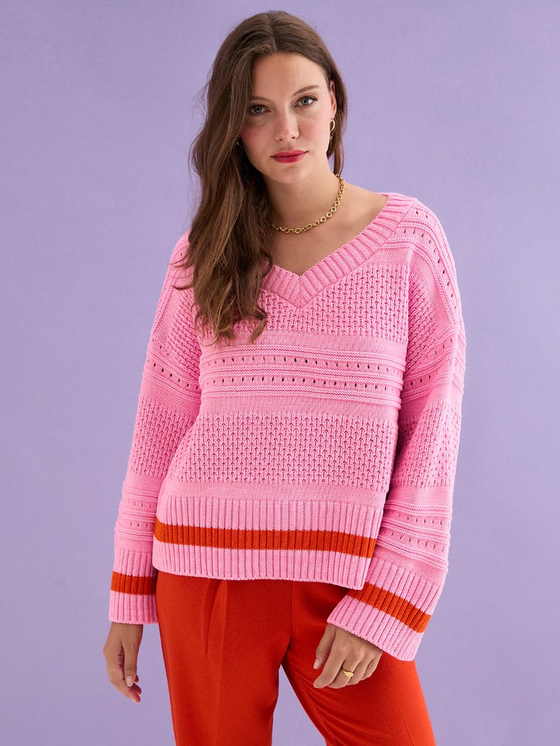 Woman wearing Cara & The Sky Judith Jumper  a pink sweater with red accents against a purple background