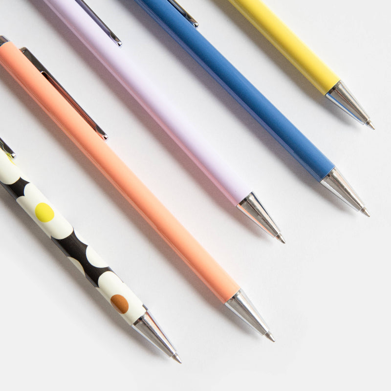 Five pastel-coloured retractable pens arranged diagonally on a clean white background. The pens are in peach, lavender, blue, yellow, and a black-and-white polka dot patterned design, with silver metal tips.
