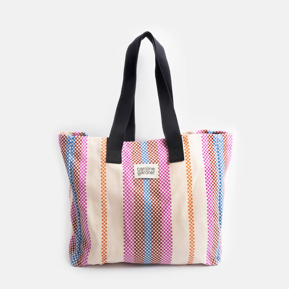 Carolien Gardner striped canvas tote bag with black shoulder straps, featuring vertical stripes in pink, orange, blue, and beige with a small white 'carolina garden' label on the front, set against a plain light gray background.
