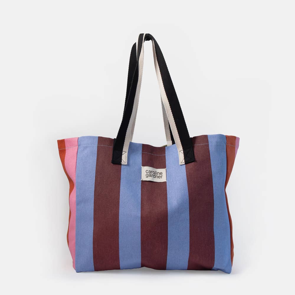 A striped canvas tote bag with vertical stripes in pink, blue, and burgundy, featuring black and white shoulder straps and a small white brand label that reads 'CAROLINE GARDNER.' on a light gray background.