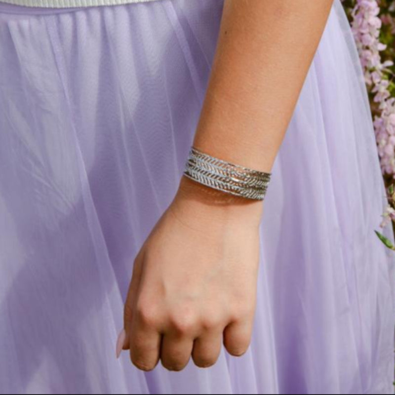 Person wearing a white top and purple skirt with a bracelet, standing in a floral setting.