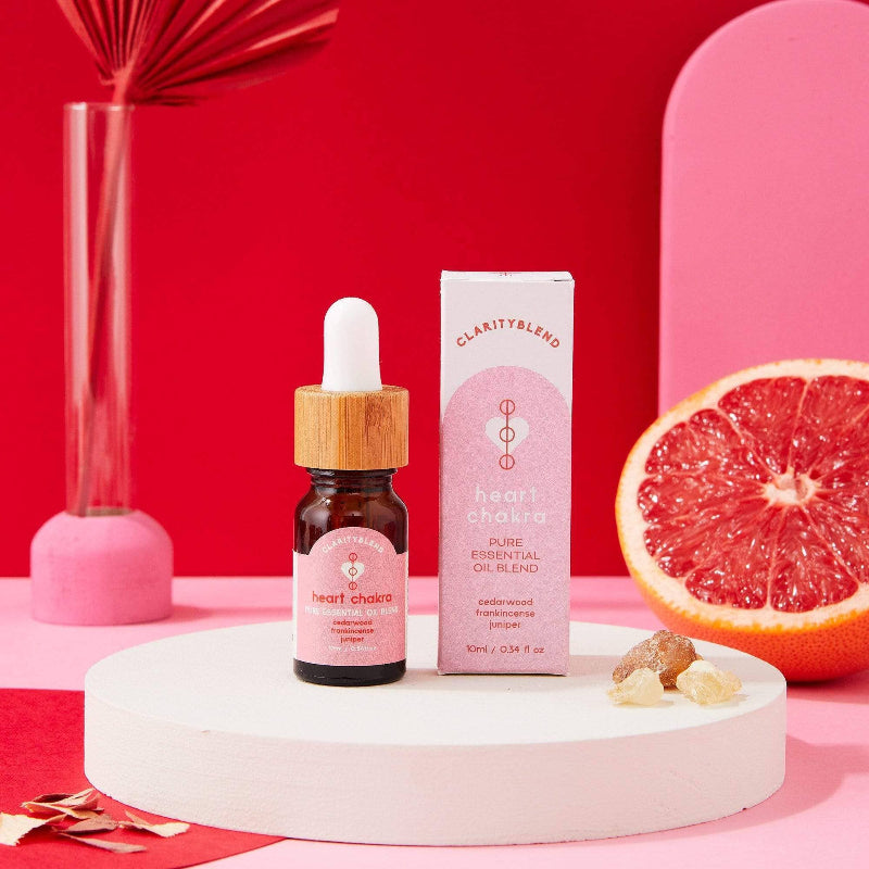 A small amber glass dropper bottle and its matching pink-and-white box labeled ClarityBlend Heart Chakra essential oil, styled on a white round pedestal with a pink and red studio background and citrus garnish.