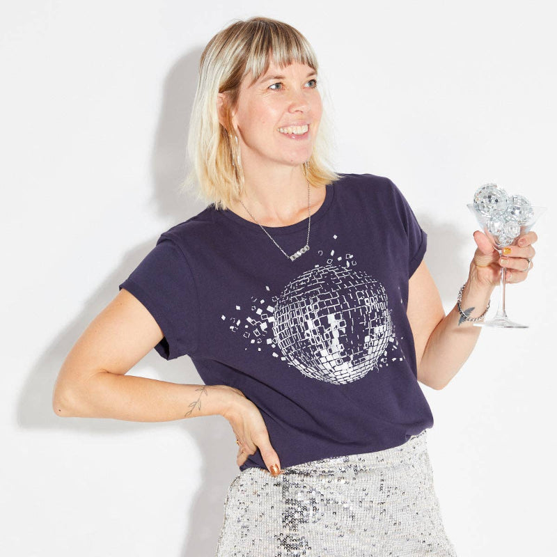 Woman wearing a disco-themed t-shirt and holding a disco ball, standing against a plain background.