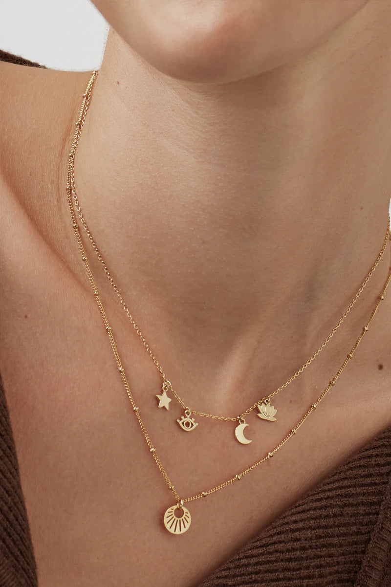 Close-up of a person wearing a gold necklace with various charms on a neutral background