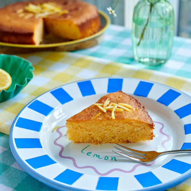 Slice of lemon cake on a plate with a fork, placed on a checkered tablecloth.