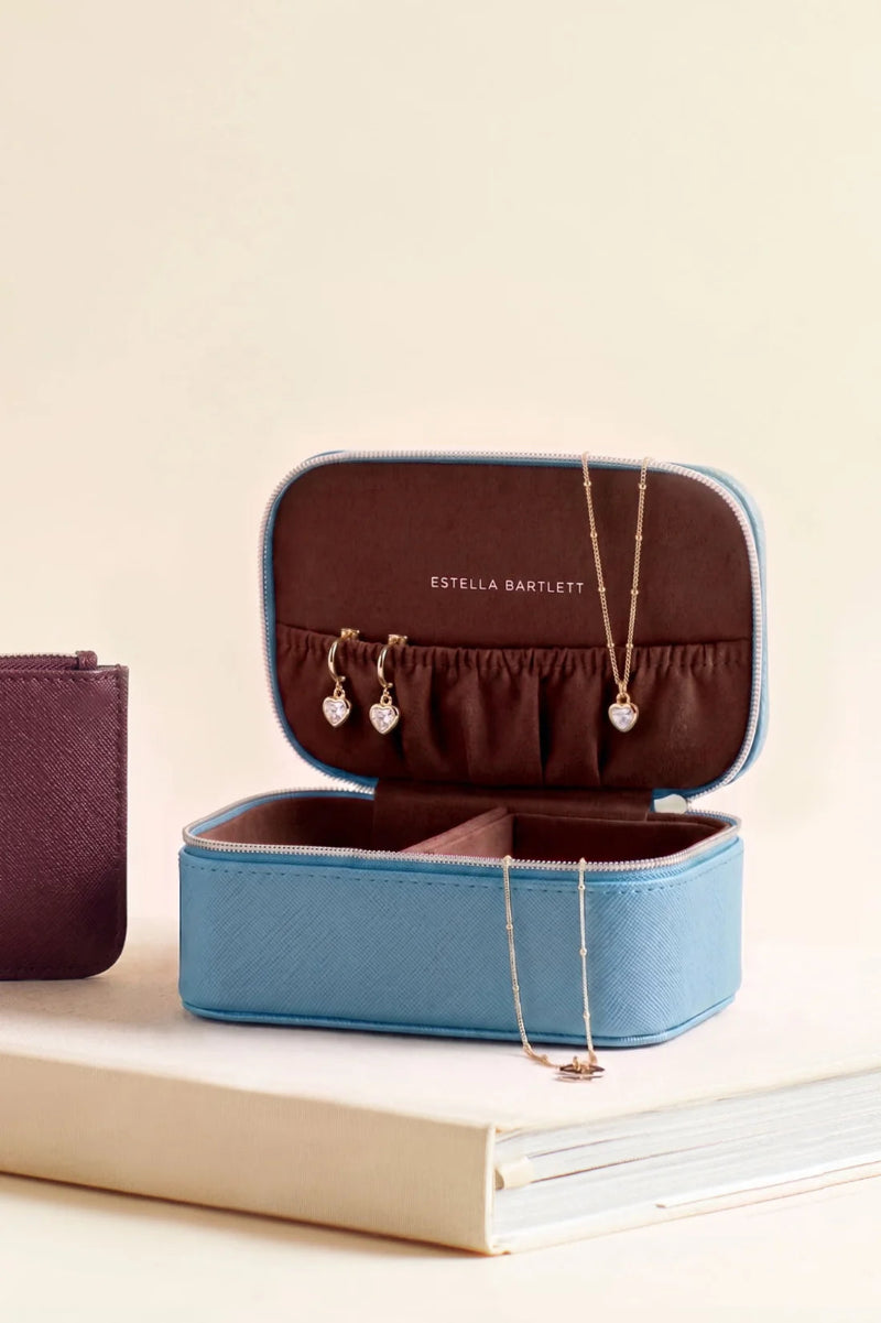 A blue and brown Estella Bartlett jewelry box, shown open with jewelry inside and draped over the rim, resting on a light-colored book against a warm cream background.